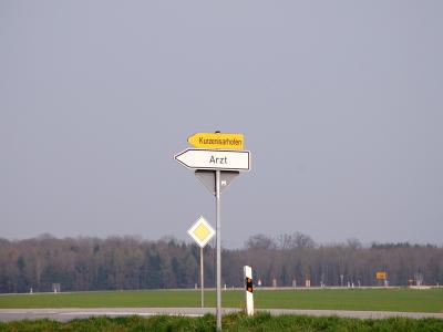 Ein Schild weist an einer Straße in auf den Weg zum nächstgelegenen Arzt hin. (Archivbild) Hinweisschild