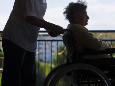 Eine Mitarbeiterin eines Seniorenheimes schiebt eine ältere Frau im Rollstuhl. Kabinett beschließt Pflege-Mindestlohn