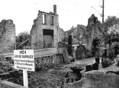 Blick auf die Ruinen von Oradour-sur-Glane (Frankreich) und eine Gedenktafel, deren Text lautet:«Gedenkstätte - Eine Gruppe von Männern wurde hier von den Deutschen massakriert und verbrannt - Besinnt Euch». Ruinen von Oradour-sur-Glane