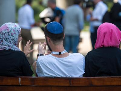 Zwei junge Muslimas mit Kopftuch und ein junger jüdischer Mann mit Kippa unterhalten sich miteinander. Begegnung