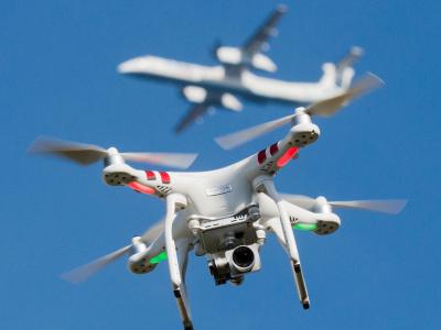 Eine private Drohne fliegt in knapp 10 Metern Flughöhe über einem Garten, in weiter Entfernung ist ein Flugzeug beim Anflug auf den Flughafen Düsseldorf zu sehen ist. Drohne und Flugzeug