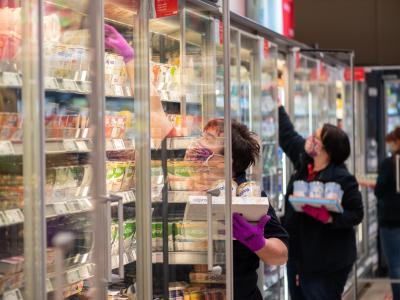 Mitarbeiterinnen in einem Supermarkt tragen Schutzmasken, während sie die Kühlregale mit Waren bestücken. Im Supermarkt